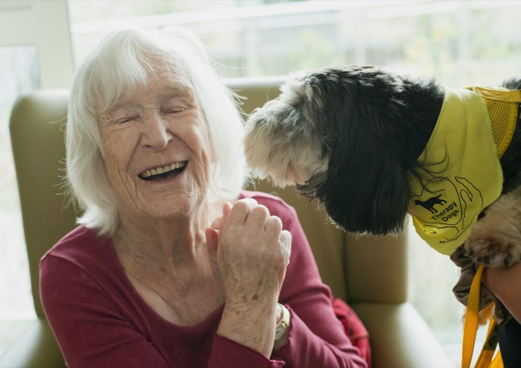 An older woman laughing as a small dog leans in towards her